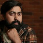 A thoughtful South Asian man with a beard, sitting indoors against a brick wall.