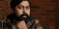 A thoughtful South Asian man with a beard, sitting indoors against a brick wall.