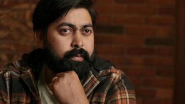 A thoughtful South Asian man with a beard, sitting indoors against a brick wall.