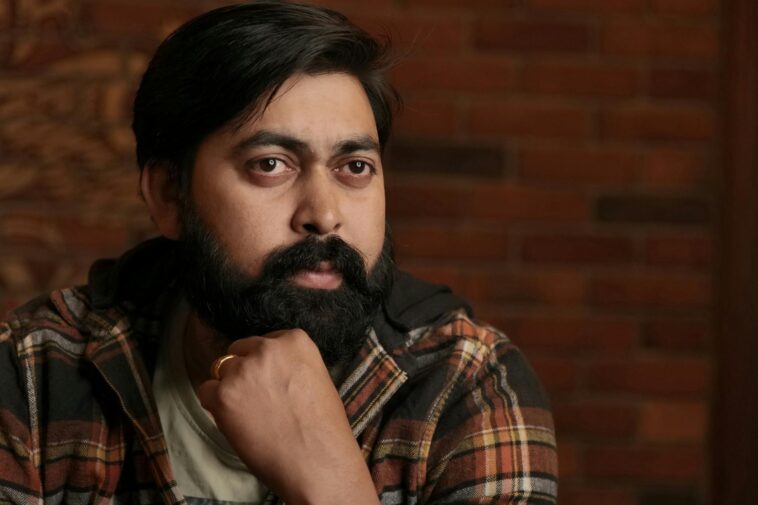 A thoughtful South Asian man with a beard, sitting indoors against a brick wall.