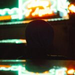 Silhouette of a person in a hoodie watching colorful neon lights at night.