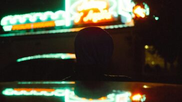 Silhouette of a person in a hoodie watching colorful neon lights at night.