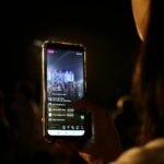 A woman live streams a concert using her smartphone at night. Vibrant stage in view.