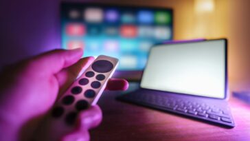 A hand using a remote to control a TV and laptop in a cozy room setting, perfect for relaxation.