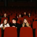 A diverse group of adults seated in a cinema, watching a movie with red seats and popcorn.