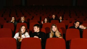 A diverse group of adults seated in a cinema, watching a movie with red seats and popcorn.