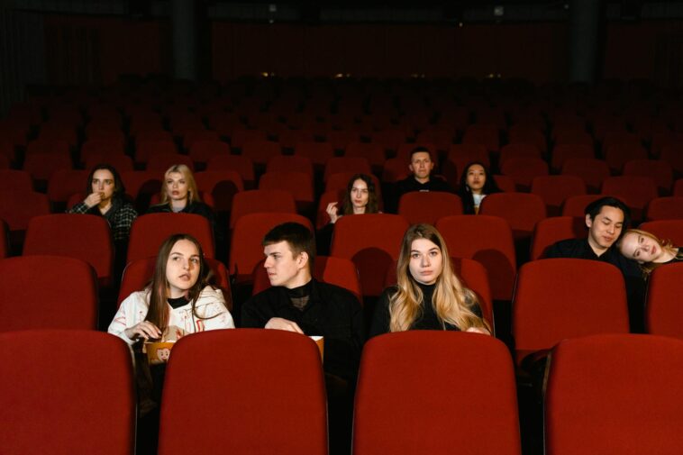 A diverse group of adults seated in a cinema, watching a movie with red seats and popcorn.