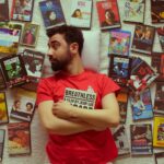 Portrait of a bearded man in a red t-shirt surrounded by vintage movie DVDs, showcasing a love for cinema.