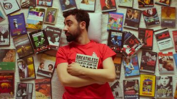Portrait of a bearded man in a red t-shirt surrounded by vintage movie DVDs, showcasing a love for cinema.