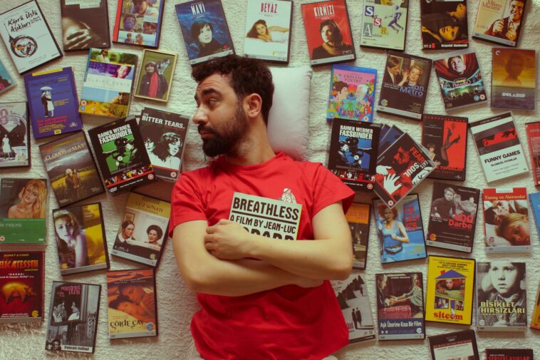 Portrait of a bearded man in a red t-shirt surrounded by vintage movie DVDs, showcasing a love for cinema.
