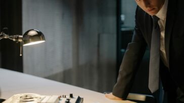 A detective in a suit examines evidence on a table under a lamp, focused on solving a case.