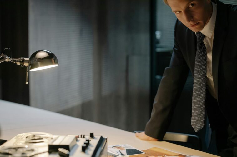 A detective in a suit examines evidence on a table under a lamp, focused on solving a case.