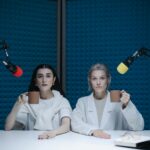 Two women in a soundproof podcast studio holding mugs next to microphones