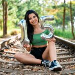 Teen girl sitting on railway tracks holding inflatable number 15 balloons, celebrating birthday outdoors.