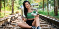 Teen girl sitting on railway tracks holding inflatable number 15 balloons, celebrating birthday outdoors.