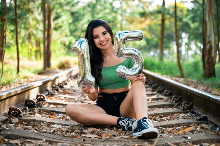 Teen girl sitting on railway tracks holding inflatable number 15 balloons, celebrating birthday outdoors.