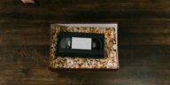 A VHS tape surrounded by popcorn in a box, placed on a wooden surface. Hands holding a marker are also visible.