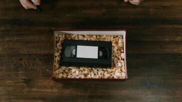 A VHS tape surrounded by popcorn in a box, placed on a wooden surface. Hands holding a marker are also visible.