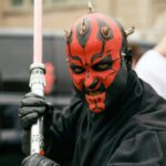 Close-up portrait of a cosplayer dressed as a Sith Lord with a lightsaber, showcasing vibrant red face paint and horns.