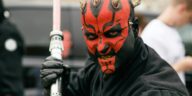Close-up portrait of a cosplayer dressed as a Sith Lord with a lightsaber, showcasing vibrant red face paint and horns.