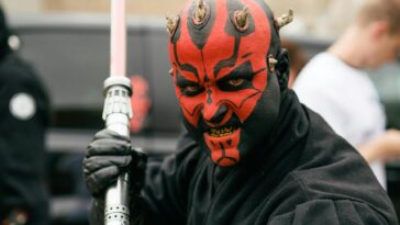 Close-up portrait of a cosplayer dressed as a Sith Lord with a lightsaber, showcasing vibrant red face paint and horns.