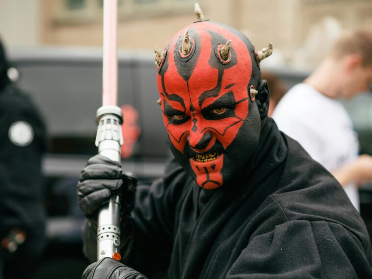 Close-up portrait of a cosplayer dressed as a Sith Lord with a lightsaber, showcasing vibrant red face paint and horns.
