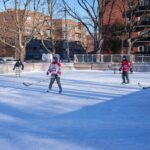 Adults playing ice hockey on an outdoor rink in a city during winter.