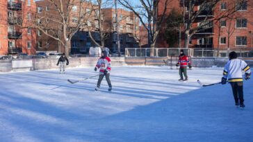Adults playing ice hockey on an outdoor rink in a city during winter.