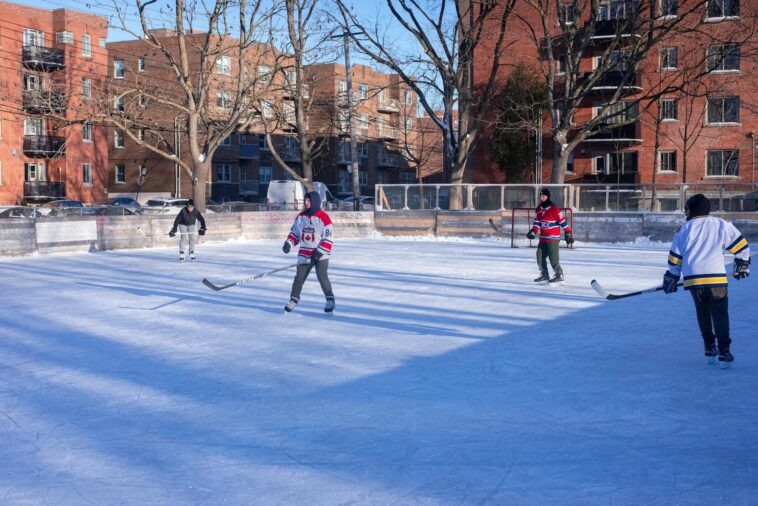 Adults playing ice hockey on an outdoor rink in a city during winter.