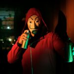 A masked person in a red hoodie holds spray paints, creating street art at night.