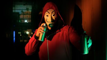 A masked person in a red hoodie holds spray paints, creating street art at night.