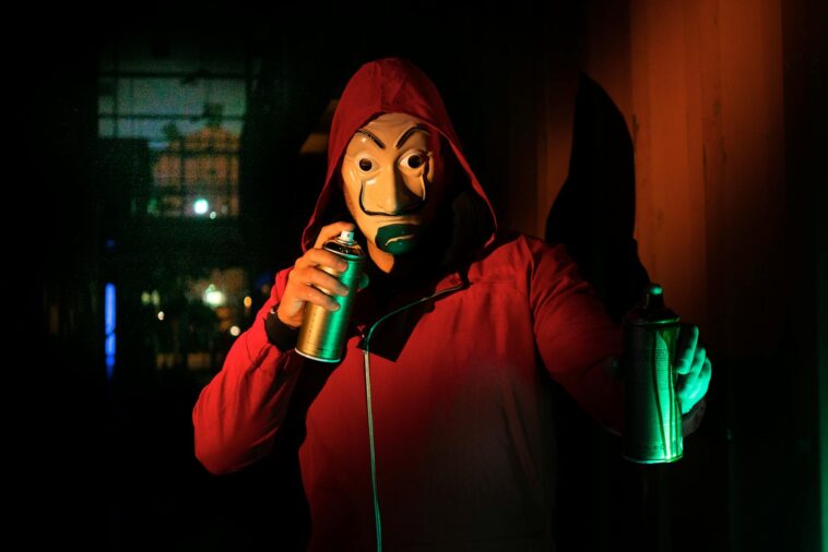 A masked person in a red hoodie holds spray paints, creating street art at night.
