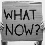A close-up of hands holding a cardboard sign with the text 'What Now?'