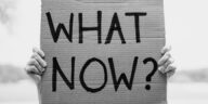 A close-up of hands holding a cardboard sign with the text 'What Now?'