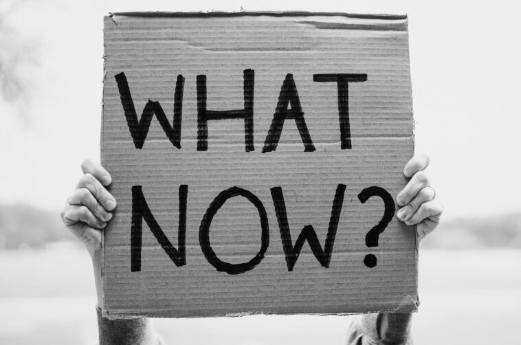 A close-up of hands holding a cardboard sign with the text 'What Now?'