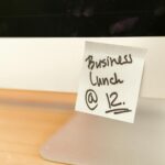 Close-up of a sticky note reminder for a business lunch at 12 o'clock on a computer monitor.