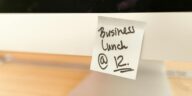 Close-up of a sticky note reminder for a business lunch at 12 o'clock on a computer monitor.