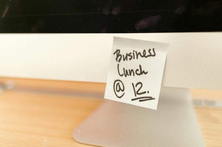 Close-up of a sticky note reminder for a business lunch at 12 o'clock on a computer monitor.