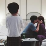 A couple shares a moment on the couch while a child stands in front, surrounded by snacks on a cozy evening.