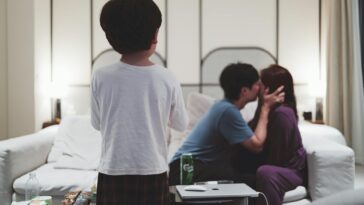 A couple shares a moment on the couch while a child stands in front, surrounded by snacks on a cozy evening.