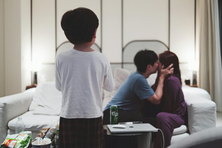A couple shares a moment on the couch while a child stands in front, surrounded by snacks on a cozy evening.