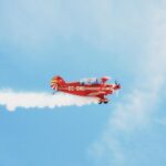A bright red biplane leaving a smoke trail against a clear blue sky.