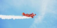 A bright red biplane leaving a smoke trail against a clear blue sky.