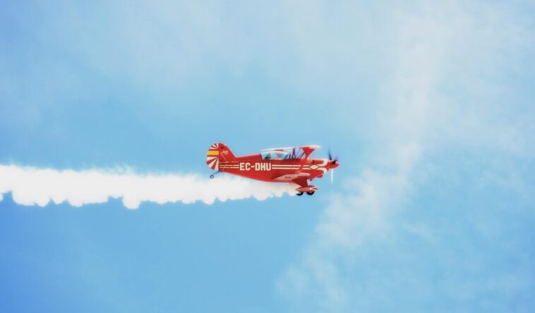 A bright red biplane leaving a smoke trail against a clear blue sky.