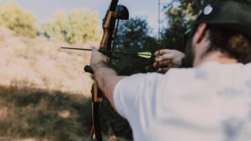 A man aiming a recurve bow during archery practice in a natural setting.