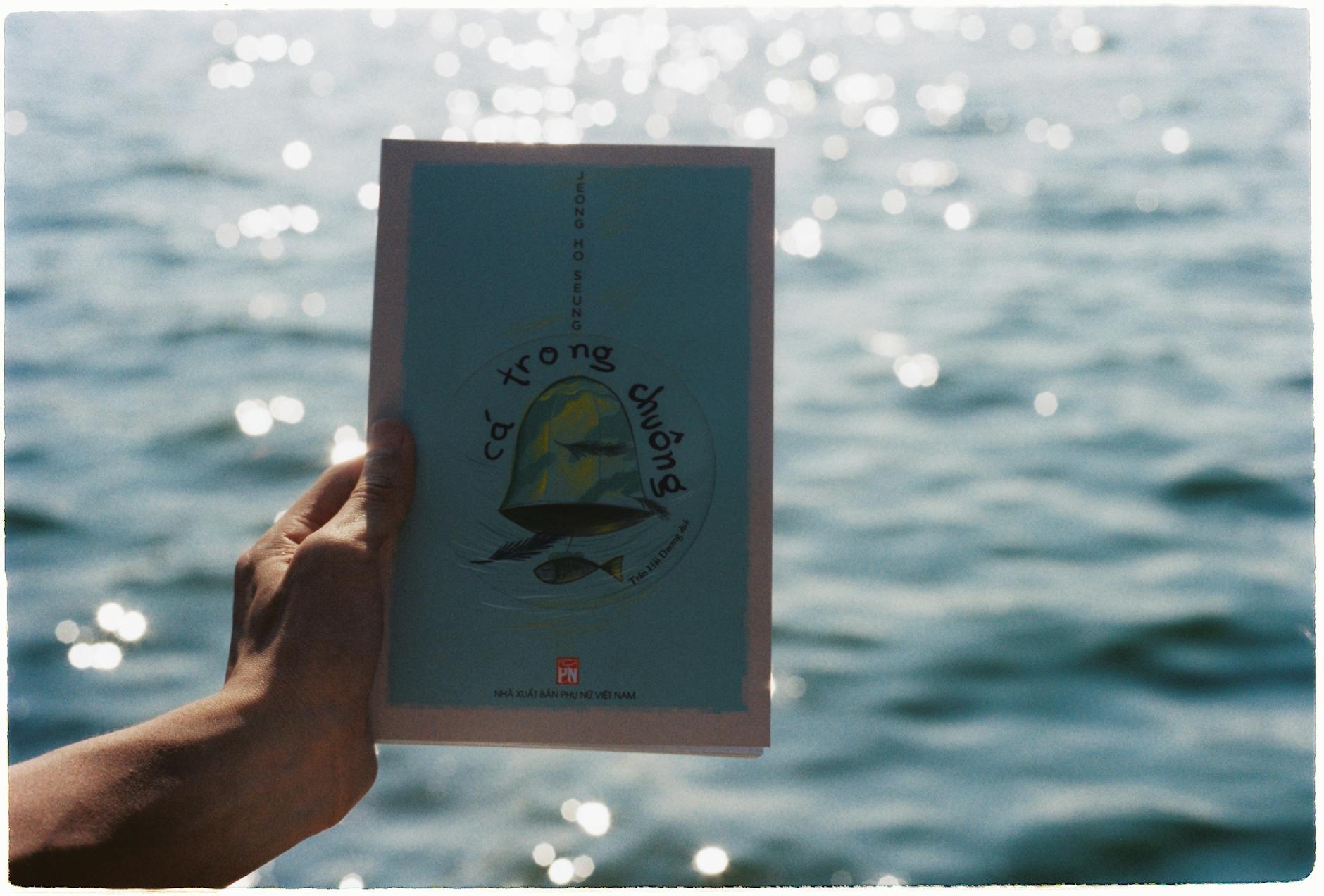 A hand holds a book against a backdrop of sparkling water, creating a calming scene.