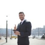 Smiling businessman in a suit giving thumbs up and holding a smartphone outdoors in a city setting.