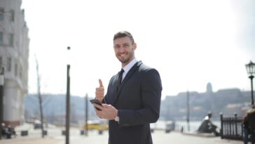 Smiling businessman in a suit giving thumbs up and holding a smartphone outdoors in a city setting.