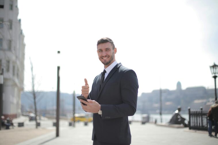 Smiling businessman in a suit giving thumbs up and holding a smartphone outdoors in a city setting.