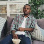 A young man relaxing on a sofa watching TV, holding a remote and popcorn.